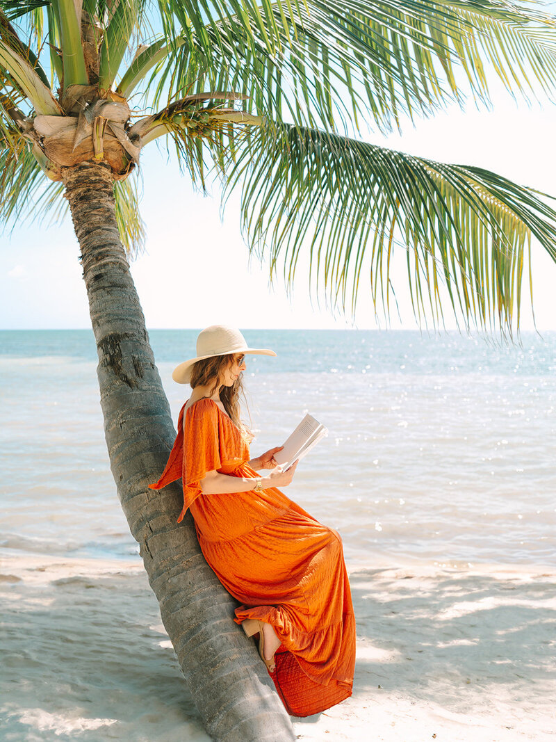 Kilee in an orange dress and straw hat sitting on a palm tree with a book in front of the ocean