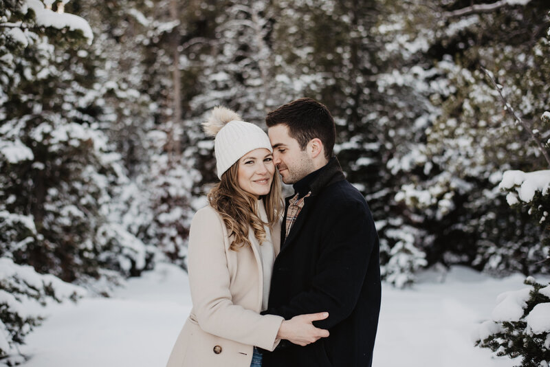 winter pines engagement pictures in the grand tetons with man in a black coat whispering into womans ear as she gently smiles