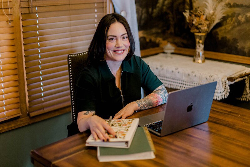 A person with shoulder-length dark hair sits at a wooden table, smiling at the camera. They have a tattooed arm resting on a book next to a laptop. Wooden blinds and a decorative vase are in the background.