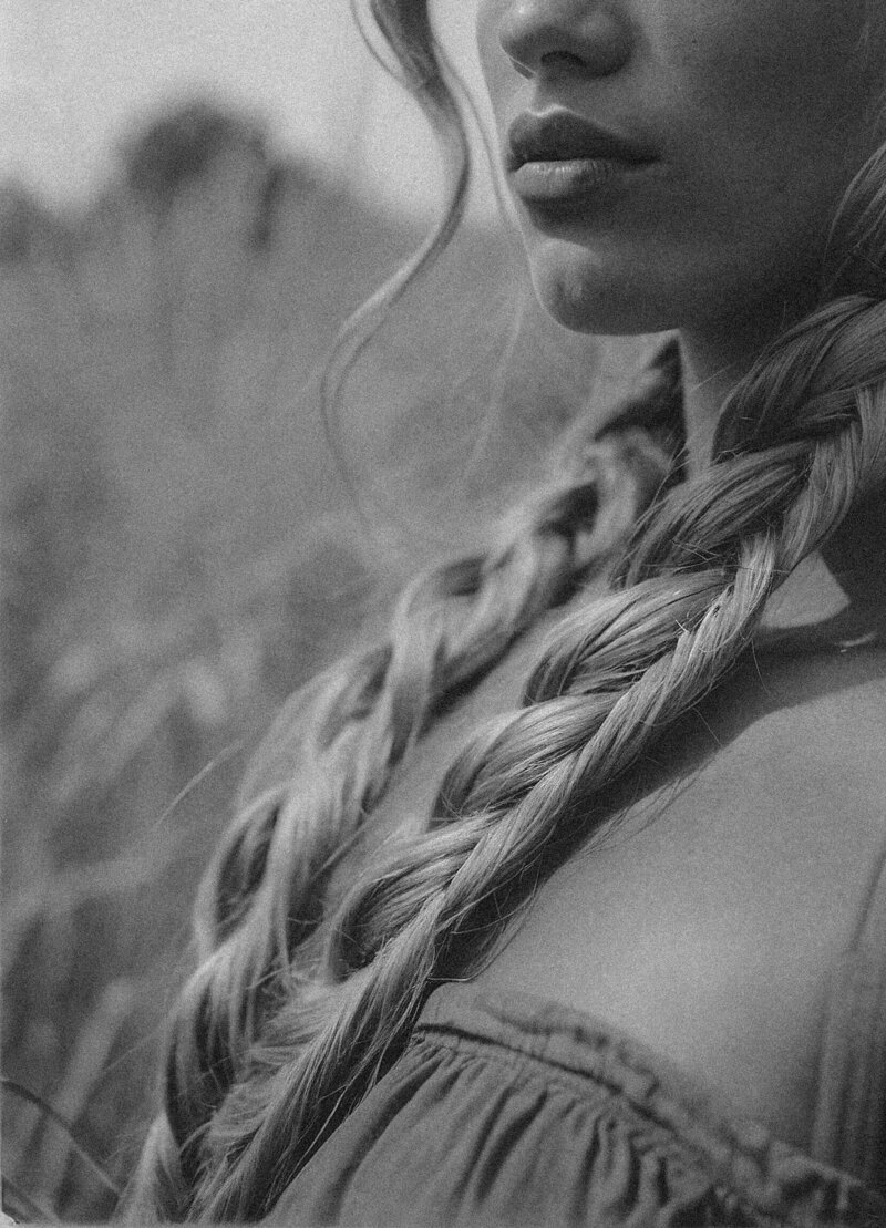 Black and white photo of a woman outdoors, showing her lower face, long braided hair, and off-the-shoulder top. The natural, blurry background evokes the artistry behind Sweet Pea Ink Creative’s unique brand design.