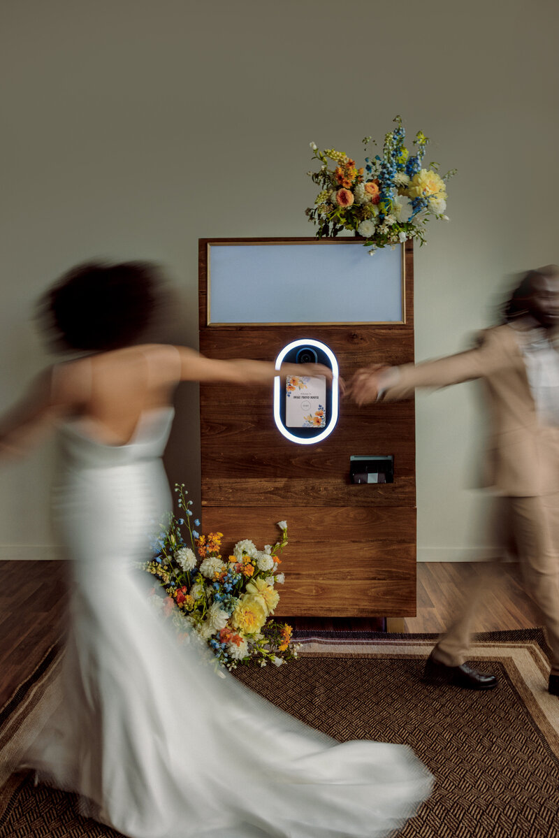 Wedding day photo booth enclosed in warm toned wood and decorated with florals while a bride and groom dance in front of it
