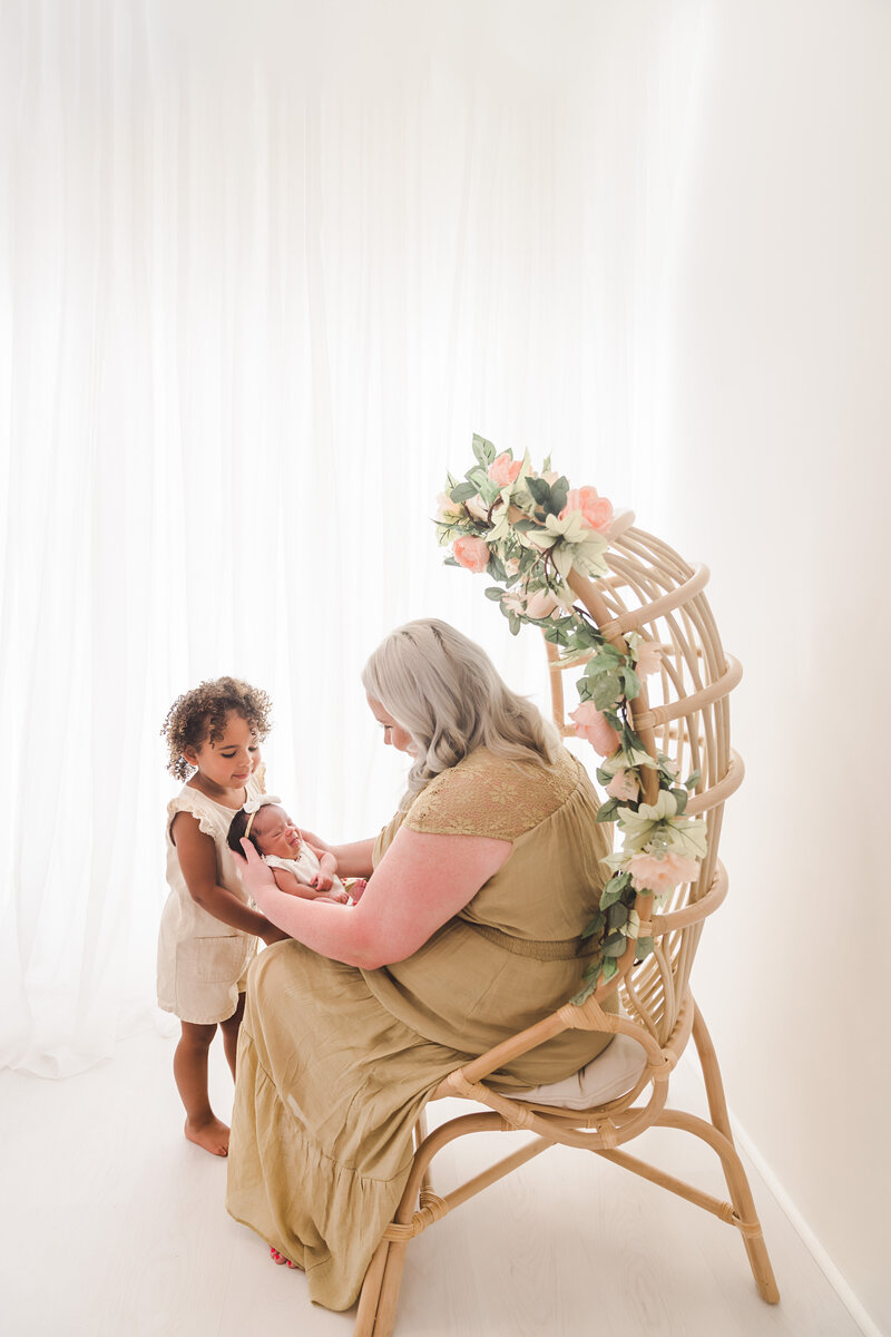 A tender moment as a mother holding a newborn interacts with her curious toddler in a softly lit room, evoking a sense of warmth, family, and love.