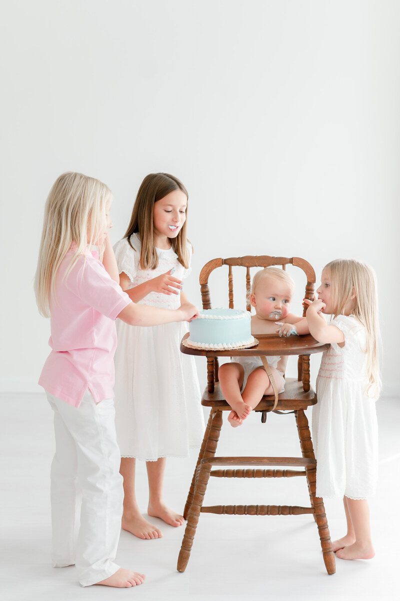 3 older siblings share a baby's cake as he sits in a high chair
