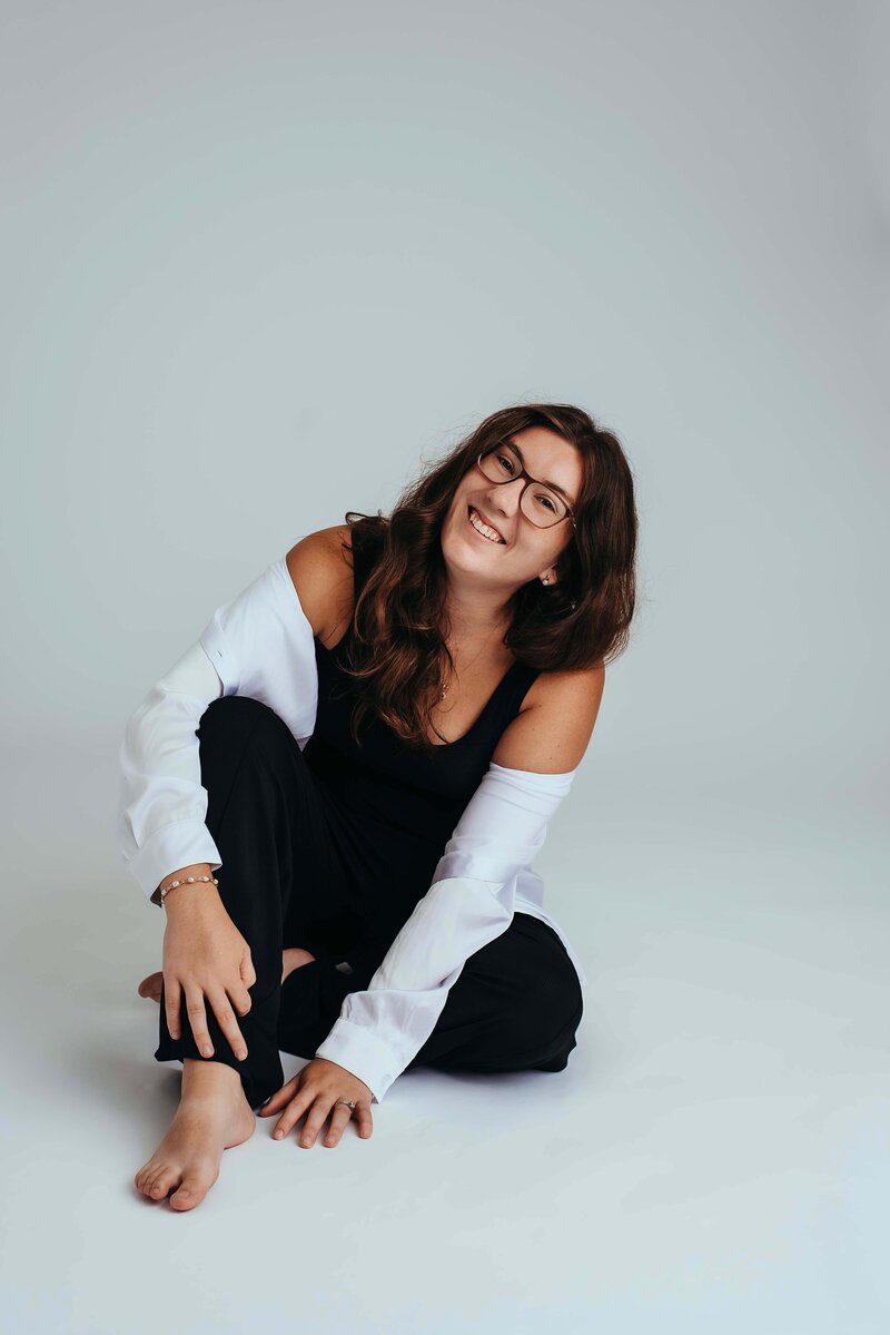 Mobile, AL newborn photographer, Molly, poses for a branding portrait. She is wearing black pants, with a black undershirt and white button up left unbuttoned. She is sitting in the floor of the studio.