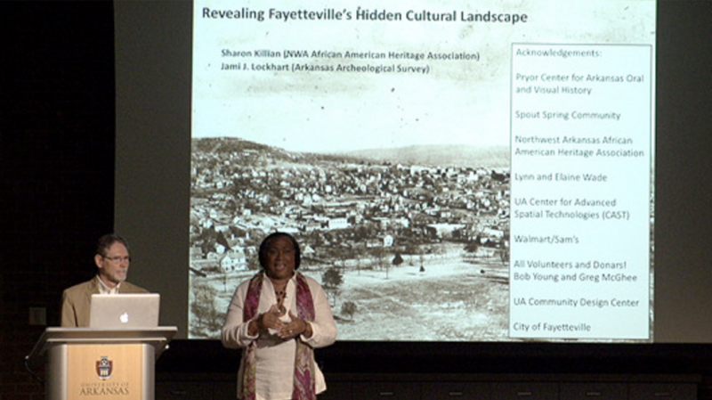 a photograph of a presentation. There are two individuals in the image: one is standing at a podium with a laptop, appearing to be presenting, and the other is standing to the side, in front of a large projection screen. The screen displays a historical photograph of a town and a title that reads "Revealing Fayetteville's Hidden Cultural Landscape" with names Sharon Killian (NWA African American Heritage Association) and Jami J. Lockhart (Arkansas Archeological Survey) mentioned presumably as speakers or contributors to the topic.