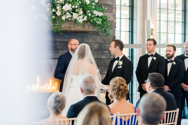 A bride and groom exchange vows in front of a romantic fireplace at Board and Batten Events, surrounded by guests in an elegant indoor wedding ceremony