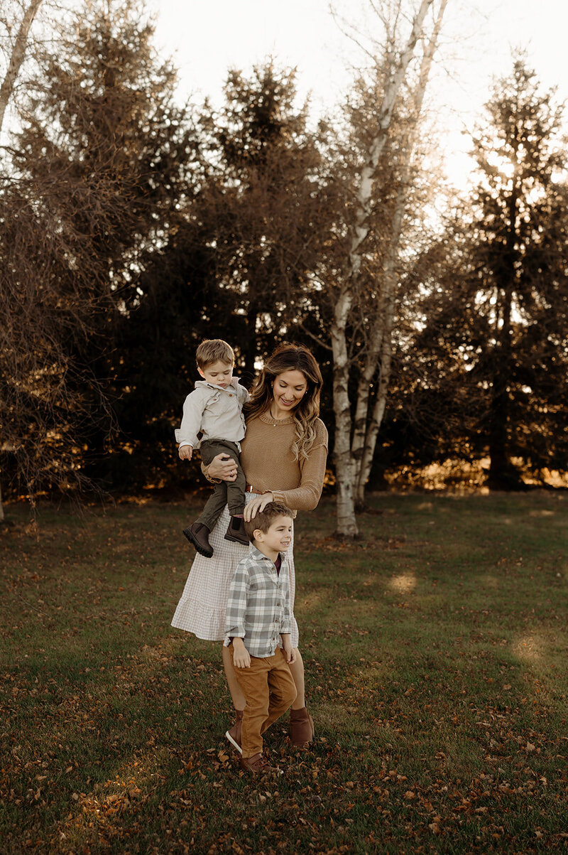 In a grassy field framed by trees, a woman stands with a young child perched on her shoulders next to another smiling child. Everyone is warmly dressed for fall, capturing the essence of New England family moments through the lens of documentary-style photography.