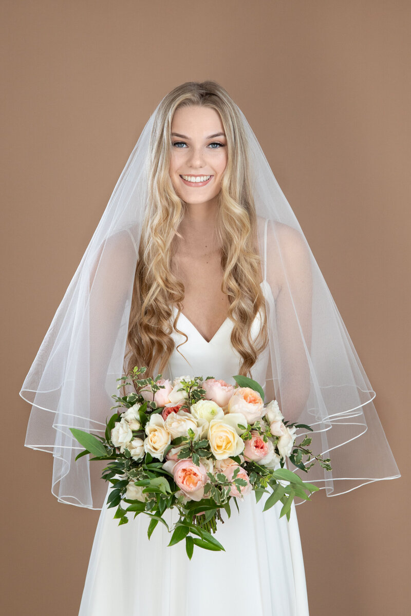 Bride wearing a fingertip length circle veil with blusher and small ribbon edge, and holding a white and blush bouquet