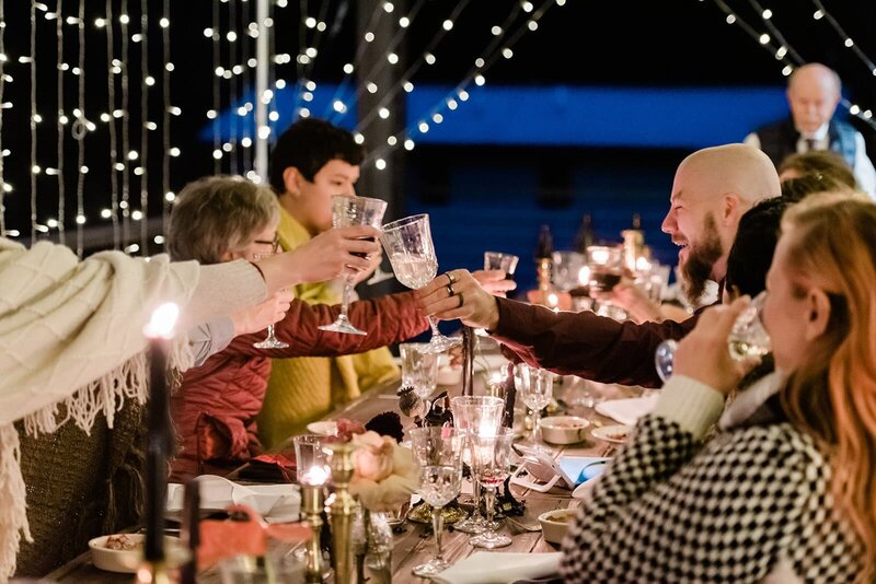 Luxury elopement photographer captures a couple sharing dinner with guests at a beautifully styled long table with candles, flowers as they toast with crystal classes, following their Alaska Glacier Elopement near Girdwood. 