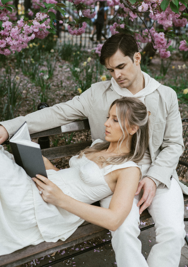 A woman leaning back on her husband sitting on a bench while reading a book