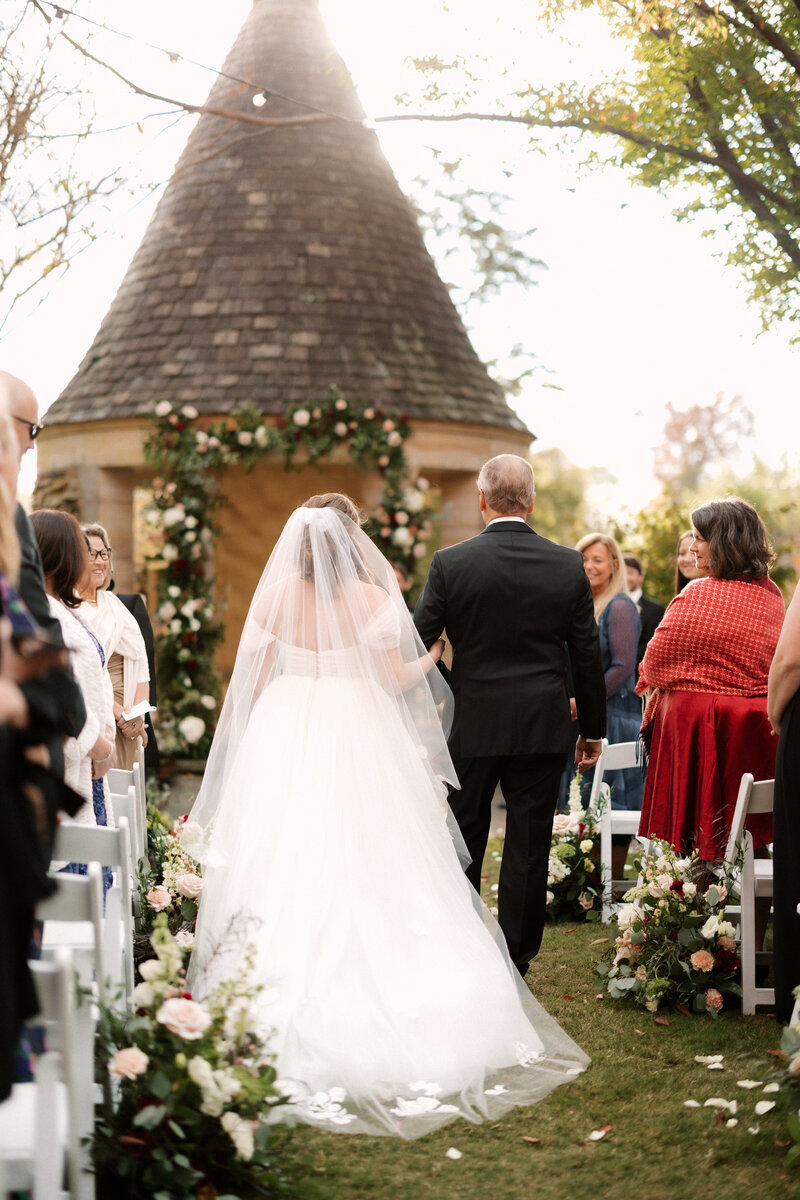 November wedding at Graylyn Estate, designed and planned by Charming Carolina — bride walking arm in arm with her father down the aisle of the White Garden ceremony space.