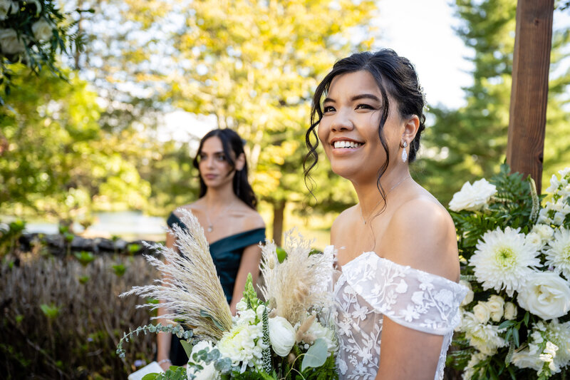laotian lesbian bride in white off the should gown standing at the ceremony alter holding flowers watching her bride come down the aisle next to a female officiant 