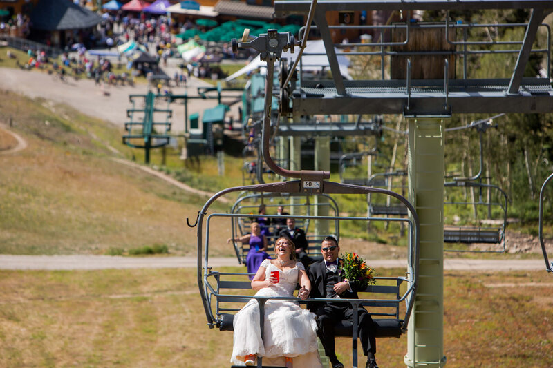 A newlywed couple rides a ski lift, the bride laughing with a drink in hand while the groom holds a bouquet. Guests follow behind, with a lively Grand Targhee resort in the background.