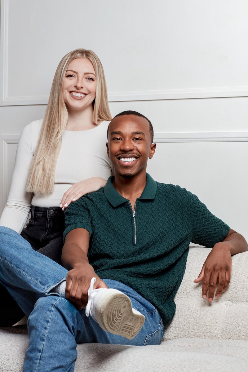 Steph and Dennis, Dennis is a young black man with short hair, and Steph is a young white woman with blonde hair. In this photo Dennis is sitting on a sofa, and Steph is sitting on the arm. They are wearing business casual clothes.