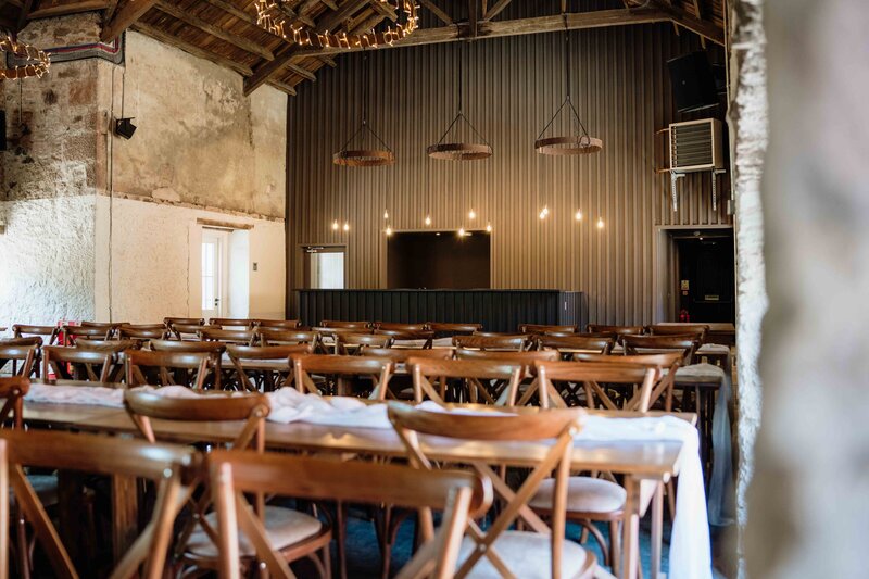 An old barn interior at Kinclune House is photographed. The space is newly renovated and is set up to host 120 wedding guests with rustic wooden chairs and tables.