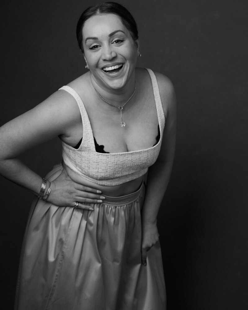 Black and white portrait of a woman mid-laugh, leaning slightly forward with joy. She wears a textured crop top with delicate straps, revealing a hint of lace, paired with a flowing satin skirt. Her accessories include a pendant necklace, bracelets, and rings, adding to her radiant confidence. A lively, unfiltered moment from 'THIS IS ME – A Photography Experience by Katie Campbell' in Atlanta, GA, celebrating self-expression and authenticity.