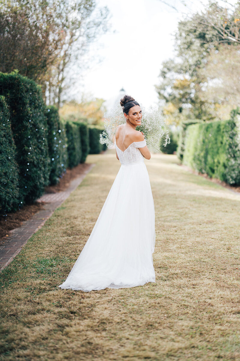 The image shows the bride in a side profile as she looks over her shoulder, giving the camera a playful smile. She is standing in a garden, her bridal gown flowing gracefully. The bride is tossing a handful of Baby's Breath into the air, which scatters around her, adding a joyful and dynamic aspect to the photo.
