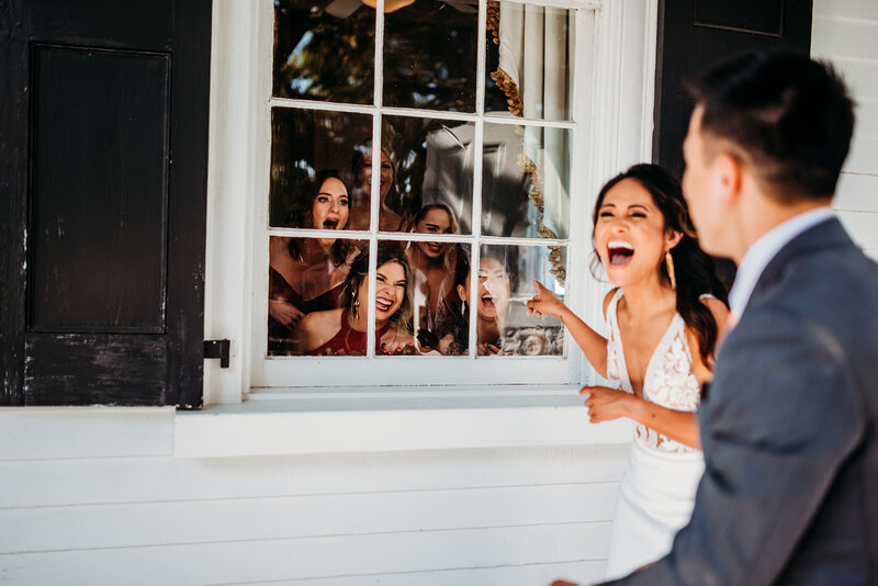 a bride and groom laughing  at their bridal party who was peaking through a window during their first look