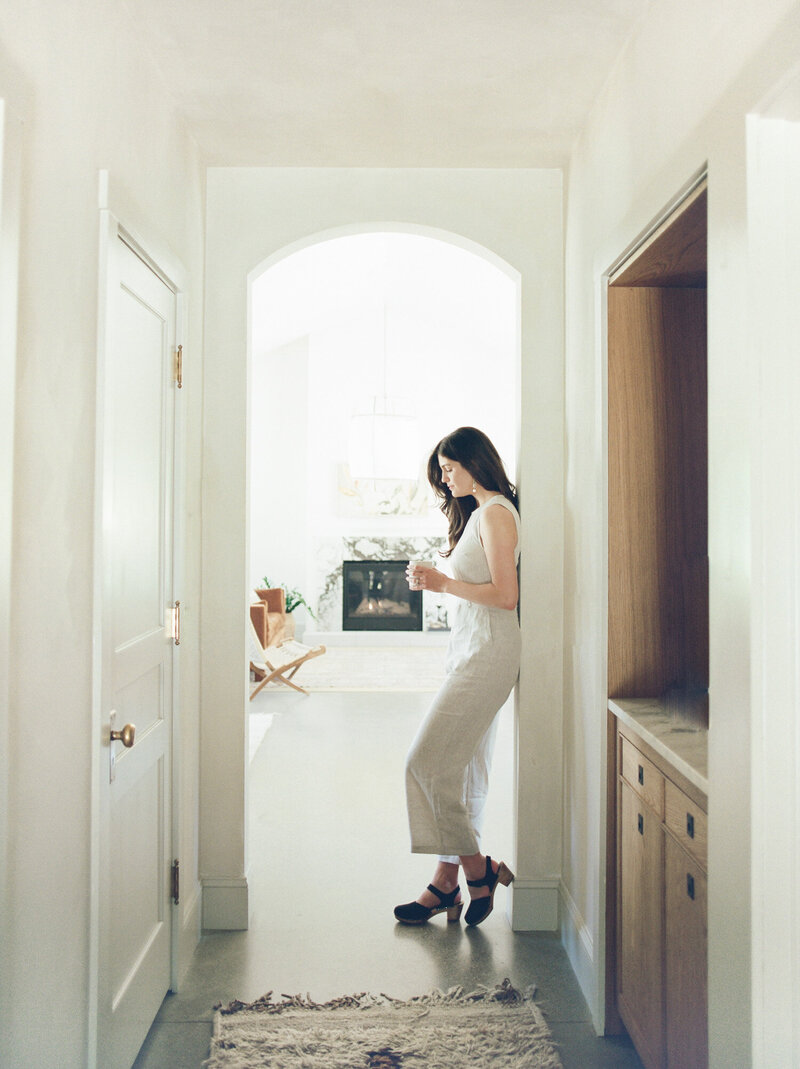 designer in linen jumpsuit relaxes with coffee, leaning in arch at Fernhaus, Northern Michigan, film branding photoshoot