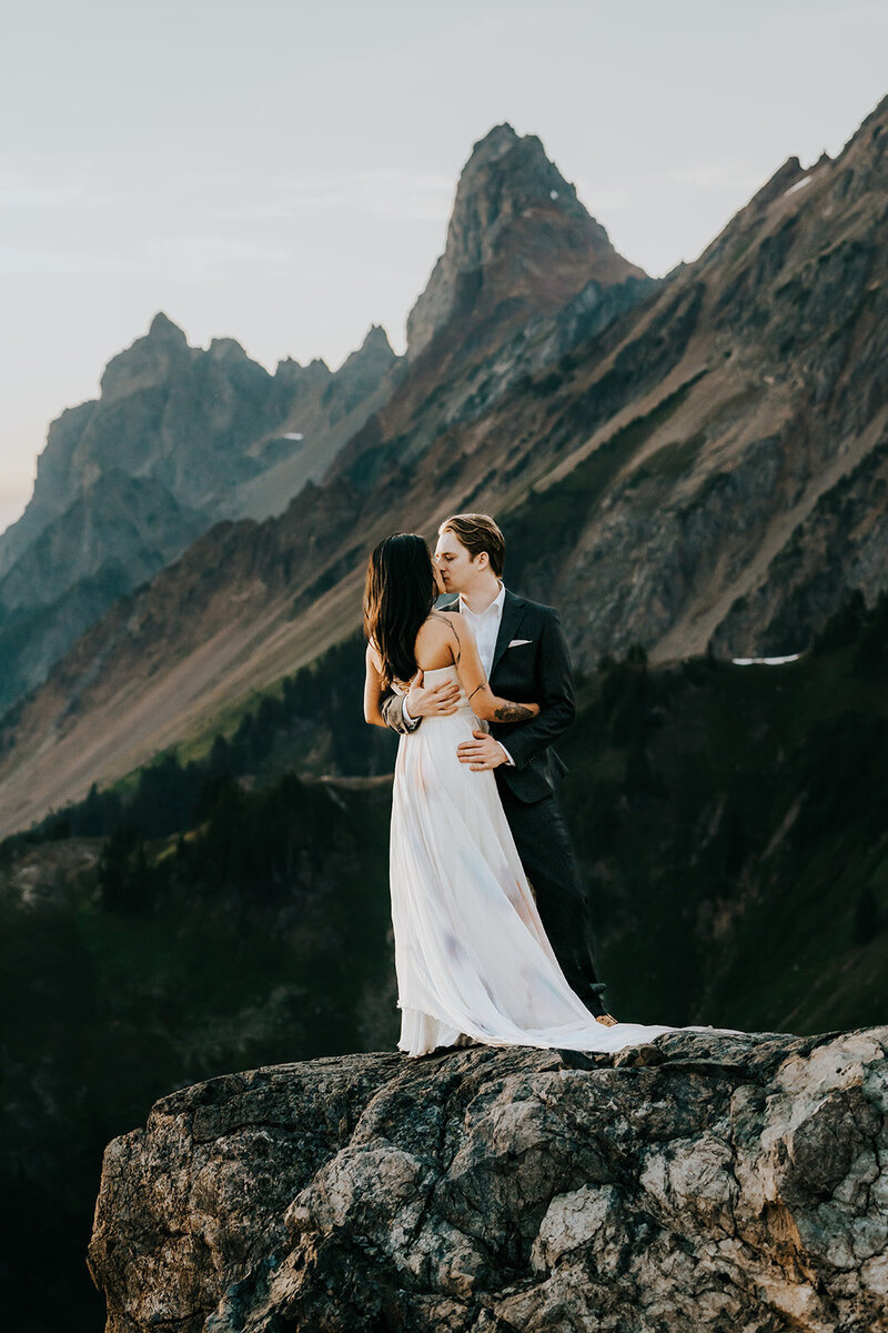 A couple in their wedding attire embrace and share a kiss as they balance on the edge of a cliff. we see the jagged mountain peaks behind them as they embrace. 