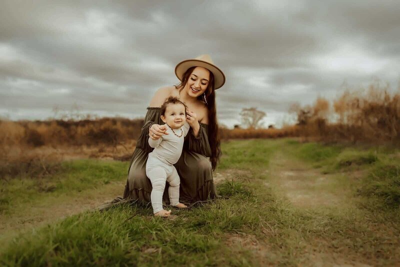 A woman in a hat kneels in a grassy field, holding a smiling baby who stands before her. With cloudy skies and a rustic landscape, this heartwarming moment is beautifully captured by a Warner Robins family photographer.