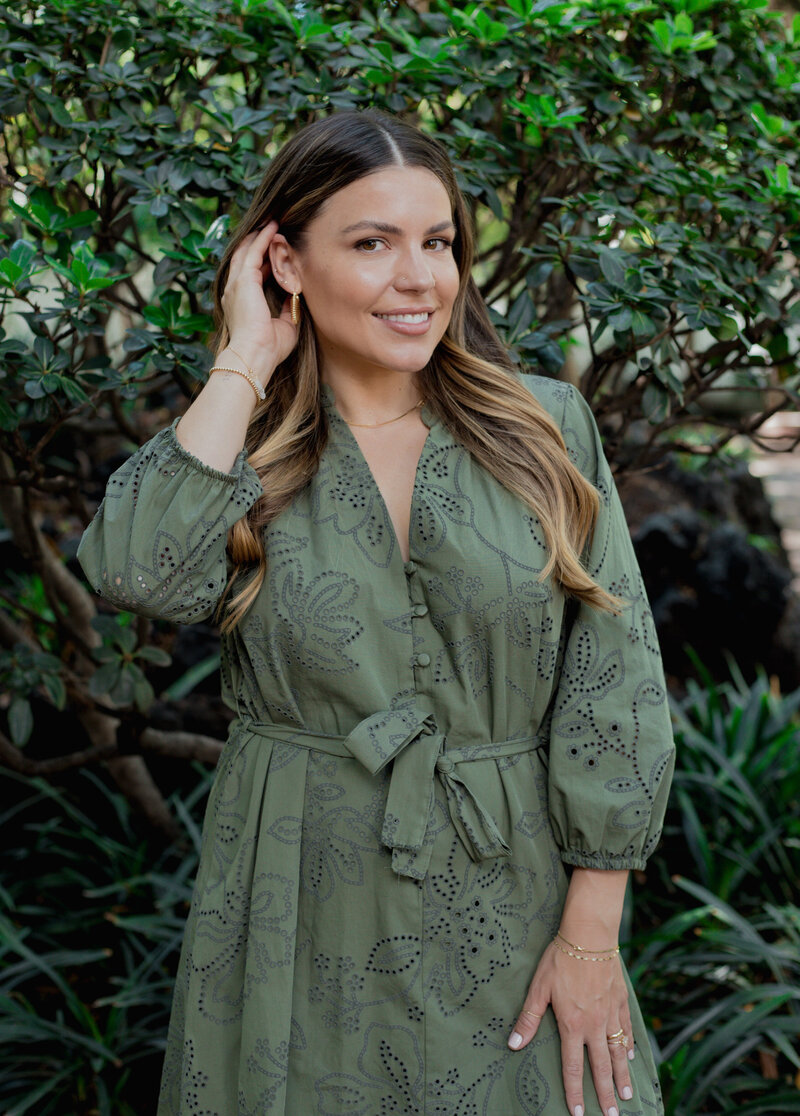Natalie Bernacchi sitting in a garden, smiling warmly in a white lace dress with greenery surrounding her on a stone path