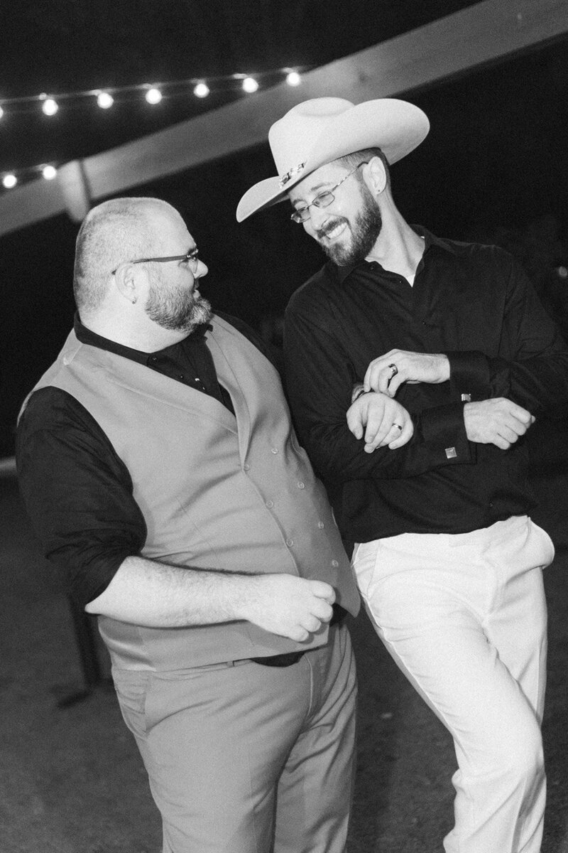 a black and white image of two men walking together as gay newlyweds in cowboy hats at their vineyard wedding venue