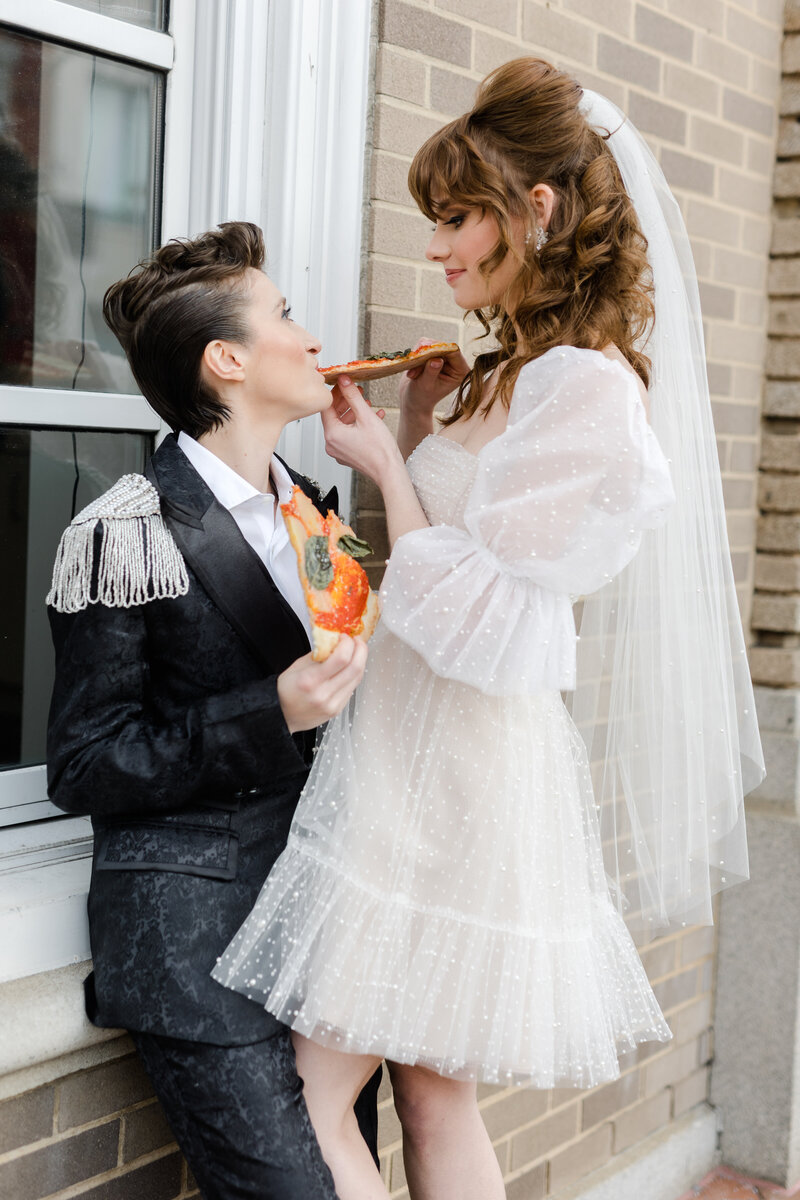 Nonbinary newlyweds holding one another leaning against a windowsill in the street of Georgetown DC feeding each other pizza 