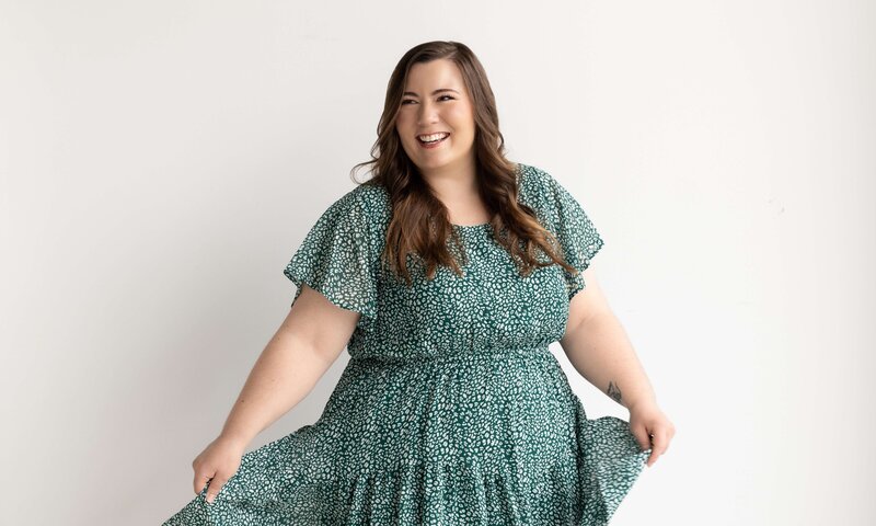 Woman in a green patterned dress smiling and twirling playfully in a bright white studio.