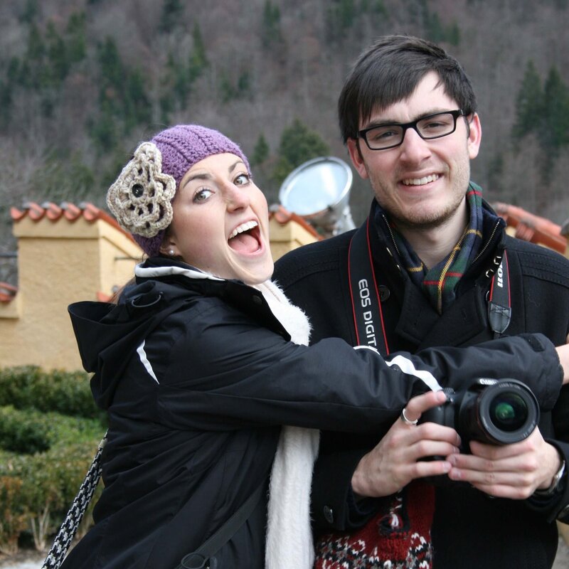 A man and a woman posing in front of a camera, woman wearing purple hat and man is holding a camera