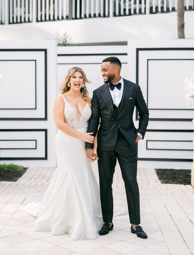Bride in wedding dress with white flower bouquet sitting in chair looking up a groom standing wearing a black tux