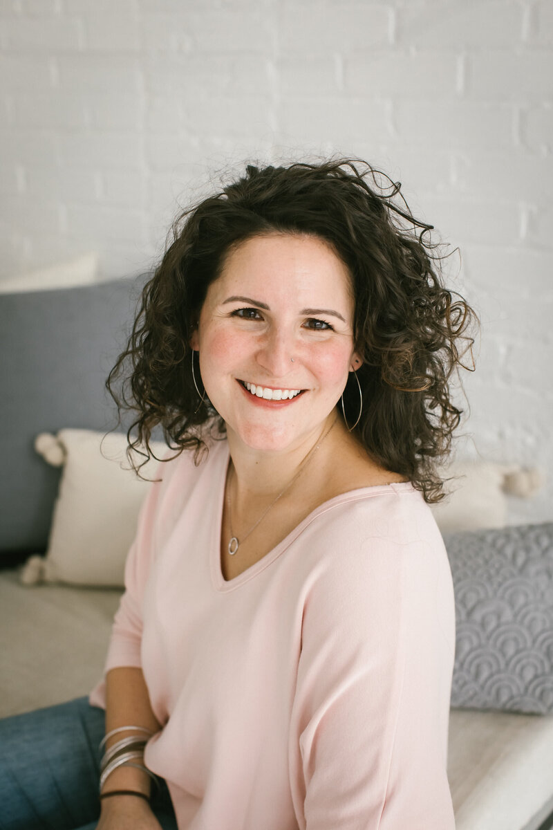 A portrait of Kate Simpson, wearing a soft pink top, sitting comfortably in a light, airy room with soft furnishings in the background.