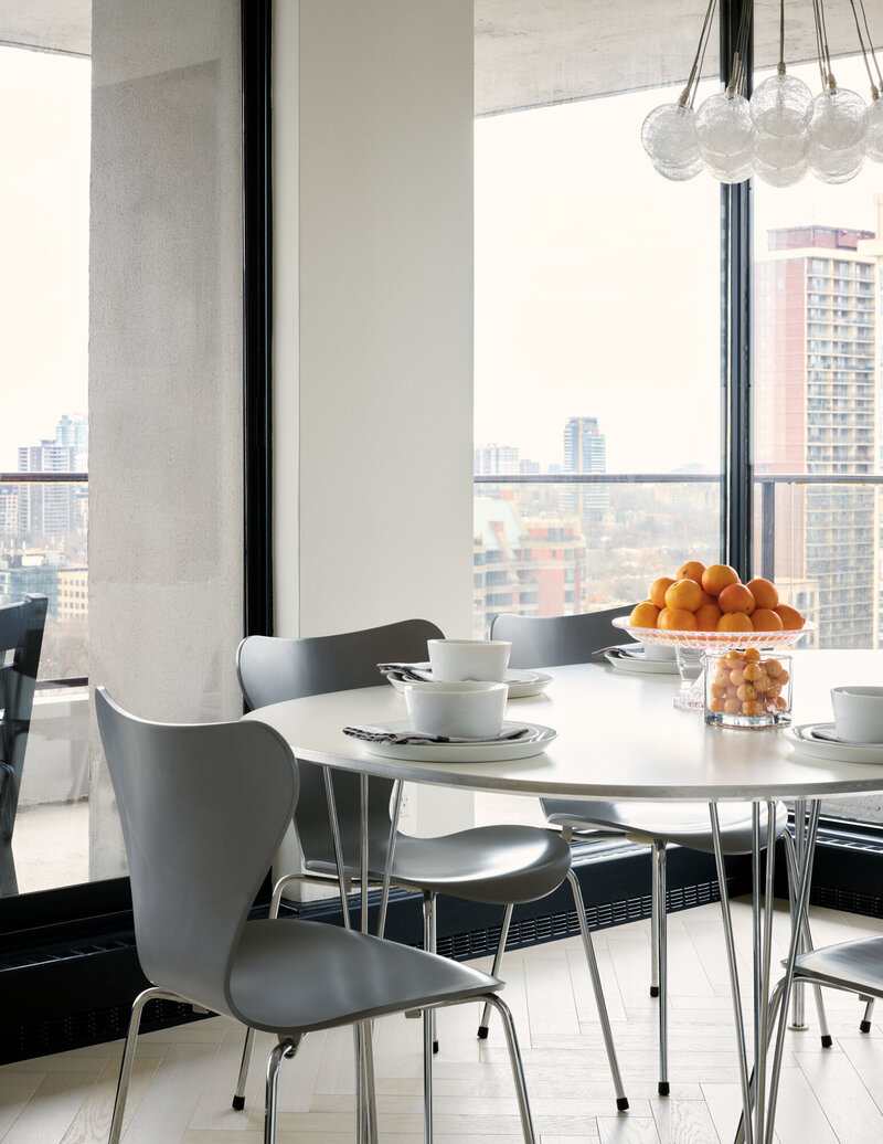 Modern, thin grey hard chairs surrounding a thin, round, white table with white bowls, plates, and a glass bowl of oranges on top of it. Large windows behind the table over look the city skyline.