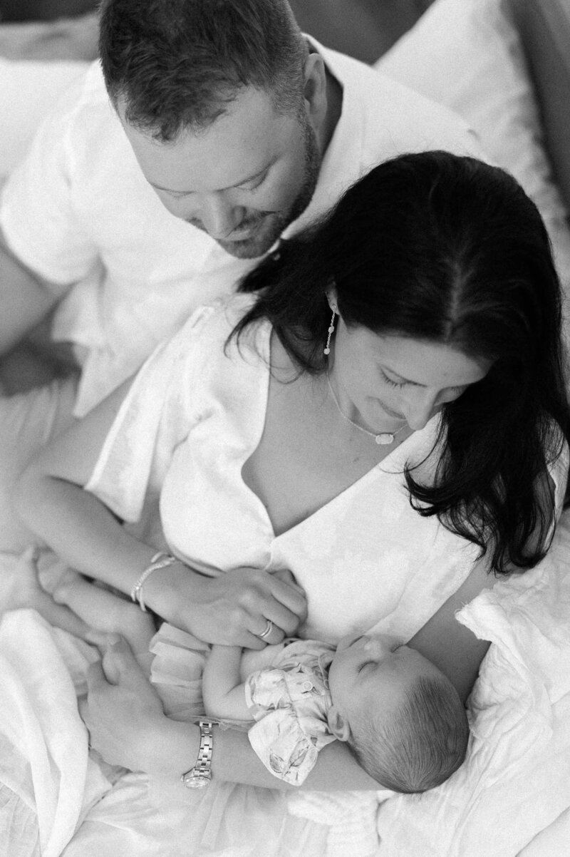 Mom and dad snuggling on a bed while holding their baby girl and looking at her lovingly while being photographed by a Los Angeles family photographer.