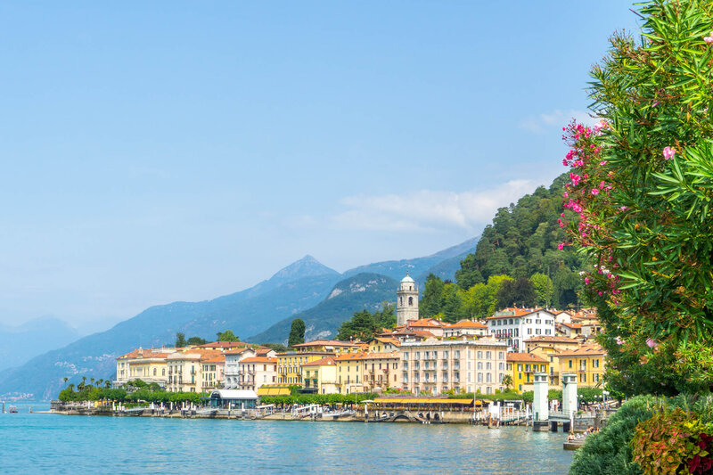A scenic lakeside town with pastel-colored buildings, a church tower, and lush green hills in the background, with bright pink flowers framing the right side of the image.