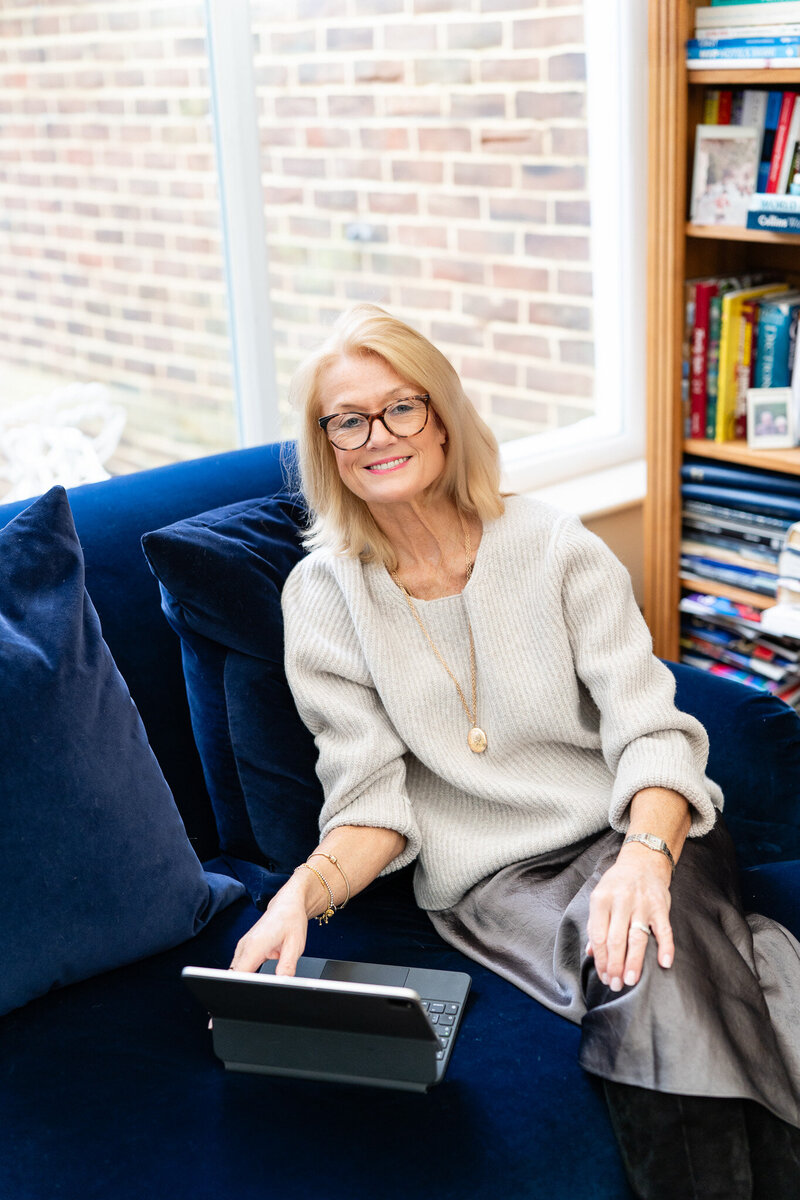woman in light beige sweather blouse sitting on a dark blue soffat with tablet in her hand and working