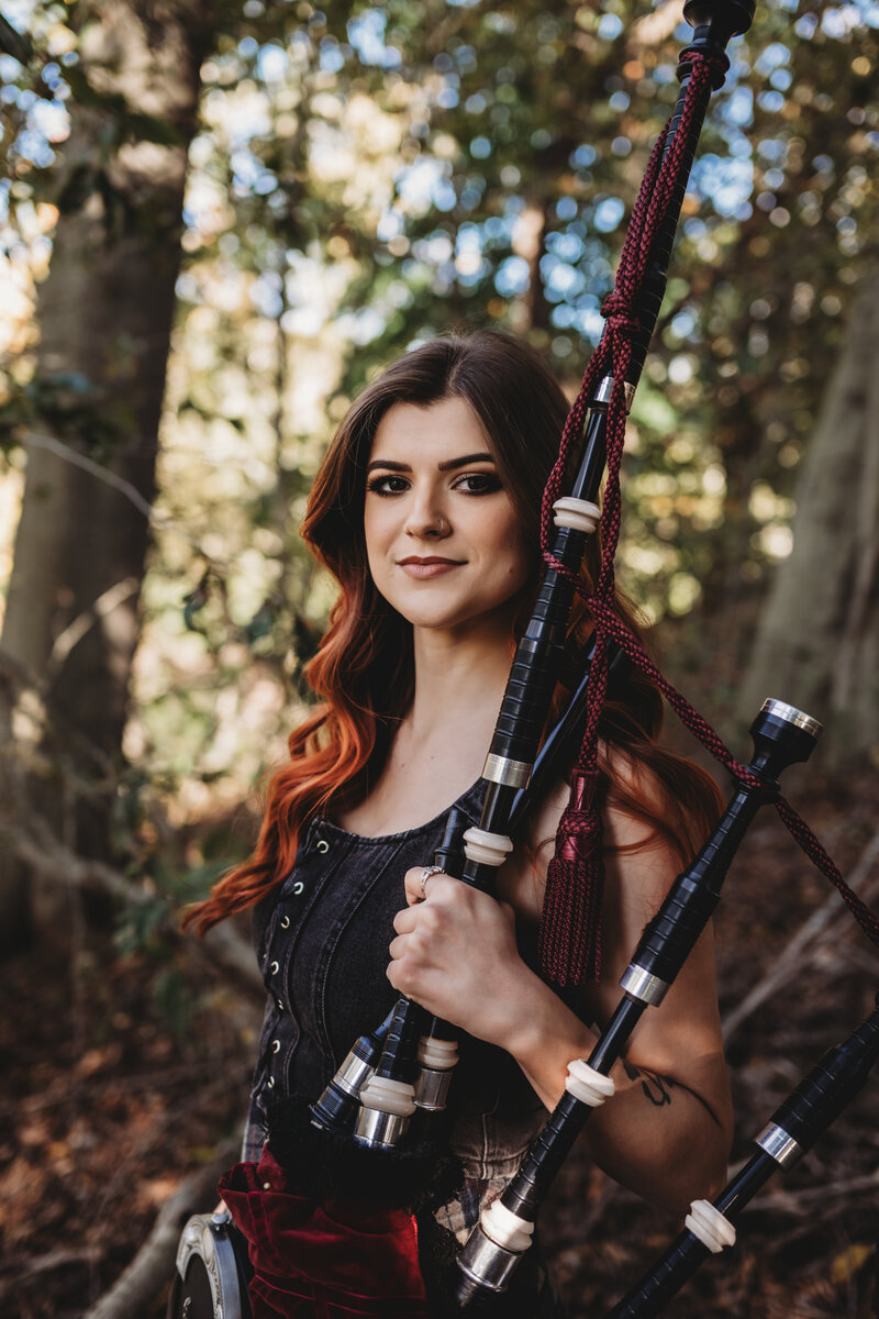 A confident young woman with long auburn hair poses in a wooded setting, holding a set of traditional bagpipes. Captured during a creative portrait photography session in Connecticut by Nicole Mele Photography, showcasing individuality and artistry.