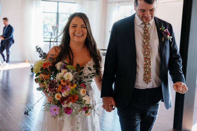 A couple in their wedding outfits hold hands, smiling