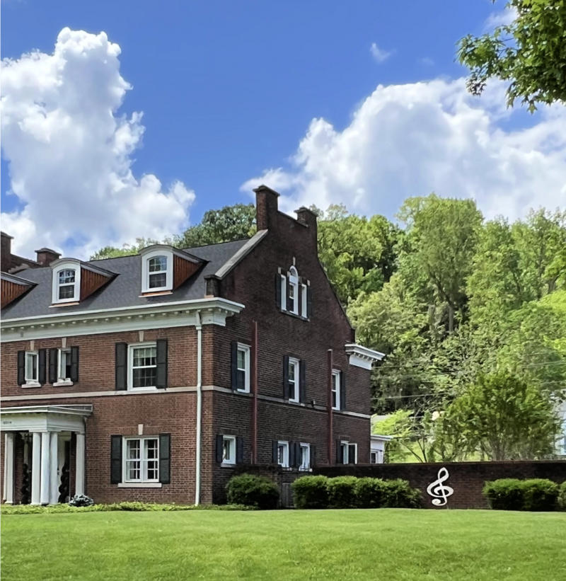 	A large brick house surrounded by green trees and a well-manicured lawn under a bright blue sky with scattered white clouds.