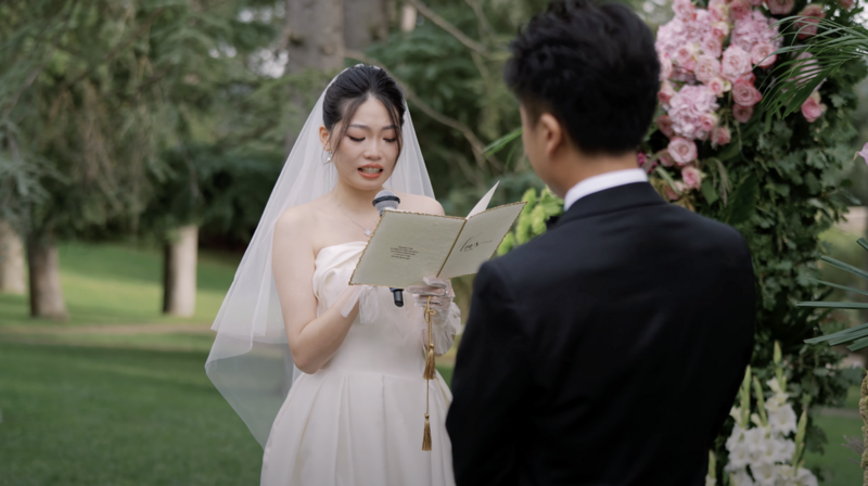 Bride reading heartfelt vows to the groom during an elegant outdoor wedding ceremony in Italy, surrounded by lush greenery and pastel floral arrangements — captured by destination wedding videographer