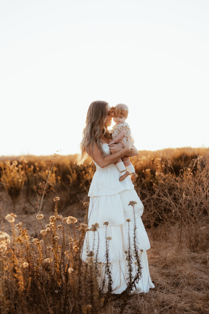 A mother in a white maxi dress holds and snuggles up to her baby girl at golden hour. 