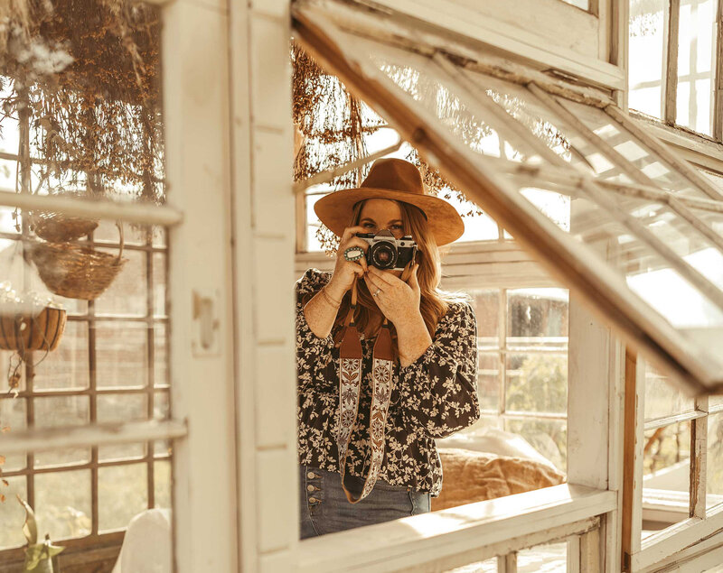 Gillian Oler the photographer holding camera in her hands  while standing inside a greenhouse. She is wearing a brown hat and a flower  top