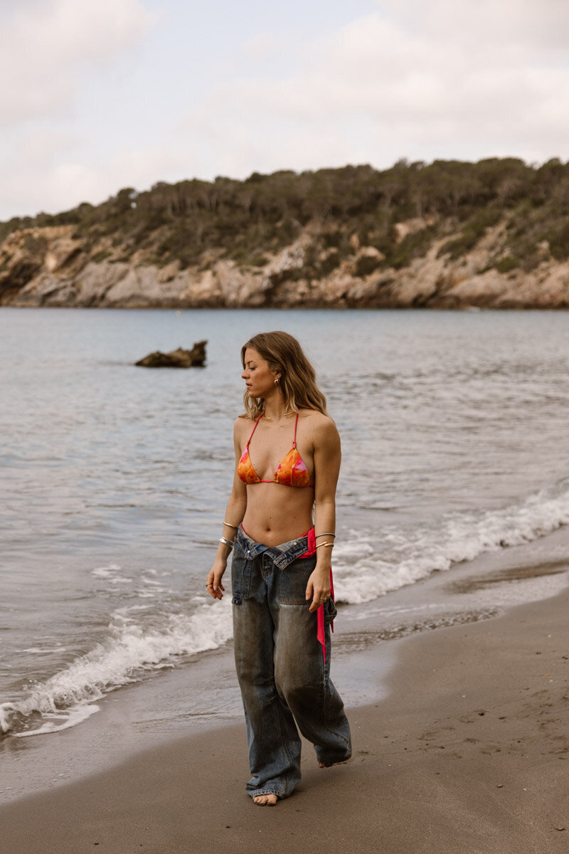 A woman walking on the beach in bikini and jeans for a branding shoot.