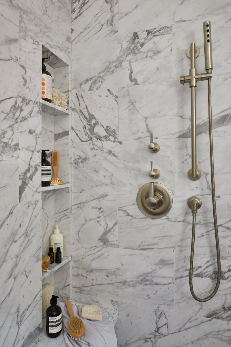 A brushed nickel shower fixture mounted to a marble wall with built-in shelves containing various soap bottles and brushes