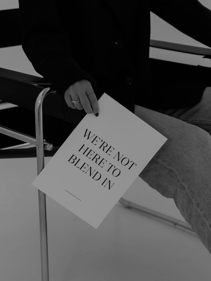 Woman seated on a modern chair holding a sign that reads “We’re not here to blend in,” symbolising bold brand presence and elevated web design.