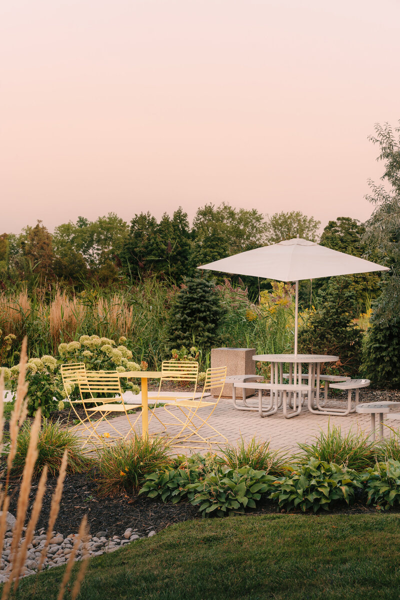 Outdoor tables and chairs with an umbrella on a patio, surrounded by lush, landscaped grounds