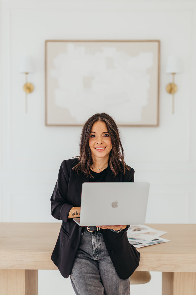 Freelance brand and web designer smiling  with a laptop in hand sitting on a messy-but-genius desk setup.