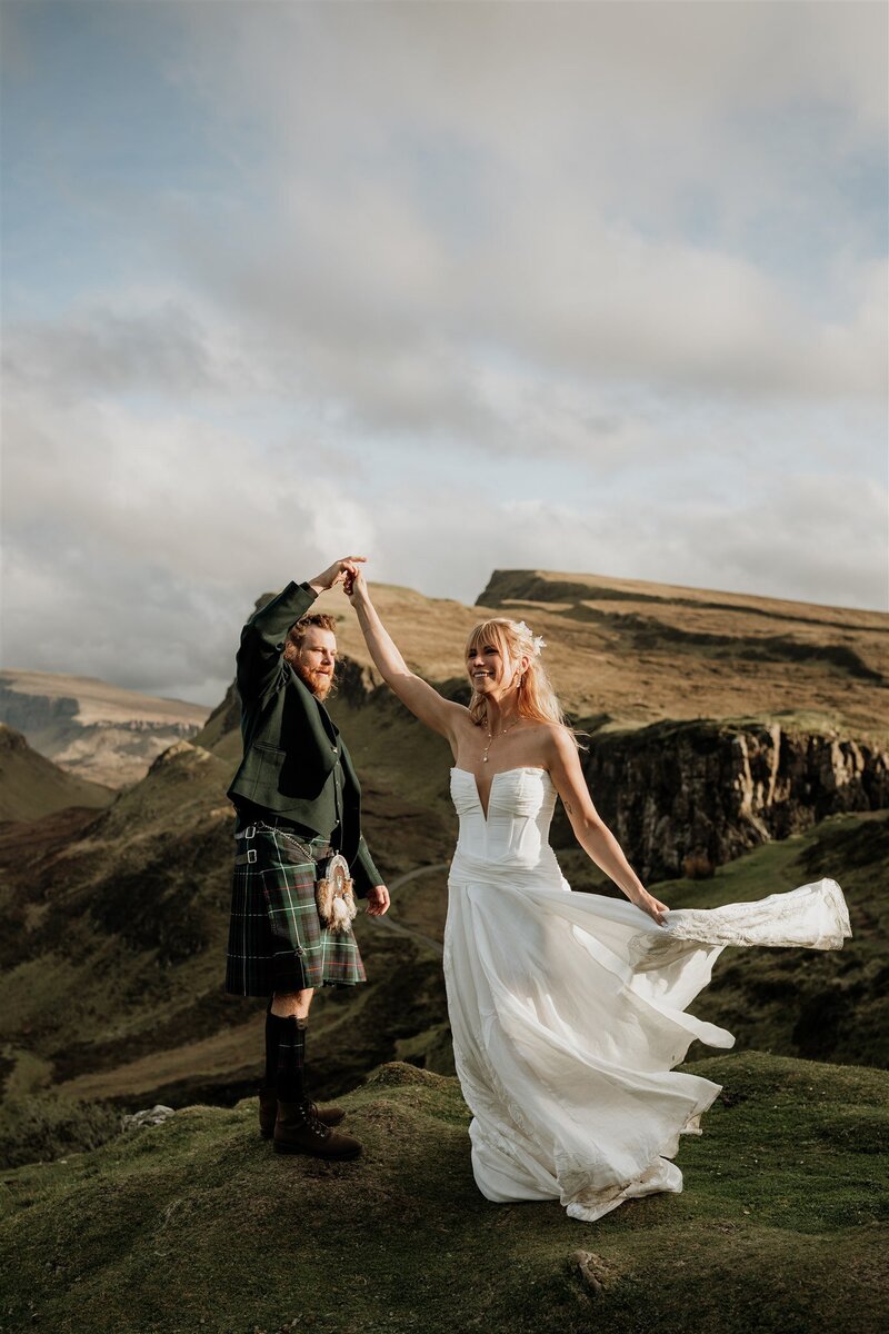 A couple are photographed on the edge of a mountain view point during their post-wedding photoshoot. They both wear wedding attire and the groom twirls the bride as the wind blows her dress. Aberdeenshire mountains are seen behind them.