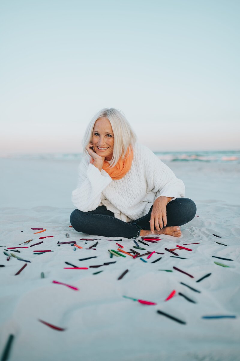 Older woman with white hair wearing a white long-sleeve shirt, jeans and an orange scarf sits cross-legged on the sand at the beach and smiles at the camera with colorful crayons strewn about surrounding her.