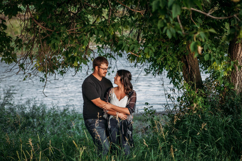 engaged couple embracing under a tree with river behind